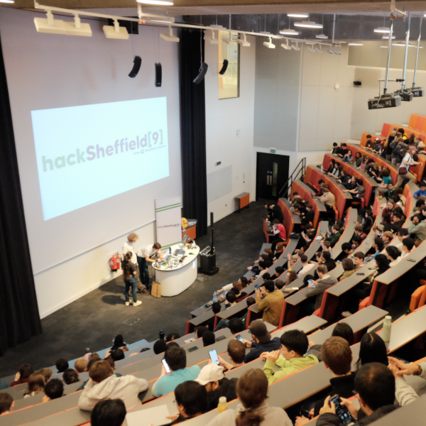 A full lecture theatre. A large screen at the front displays 'HackSheffield 9'